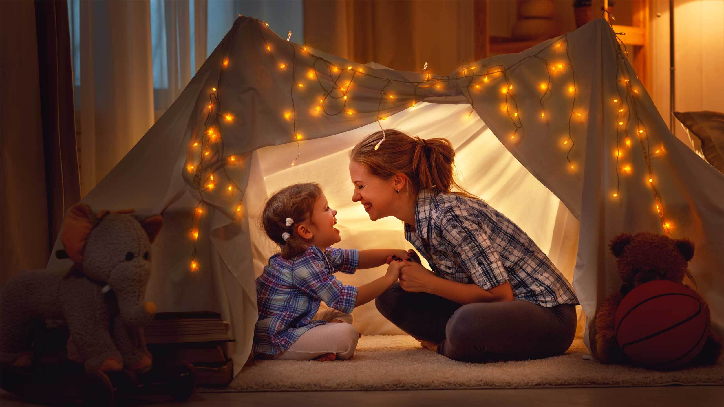 Eine Frau mit Kind in einem Zelt im Wohnzimmer bei gedämpftem Licht mit einer Lichterkette.